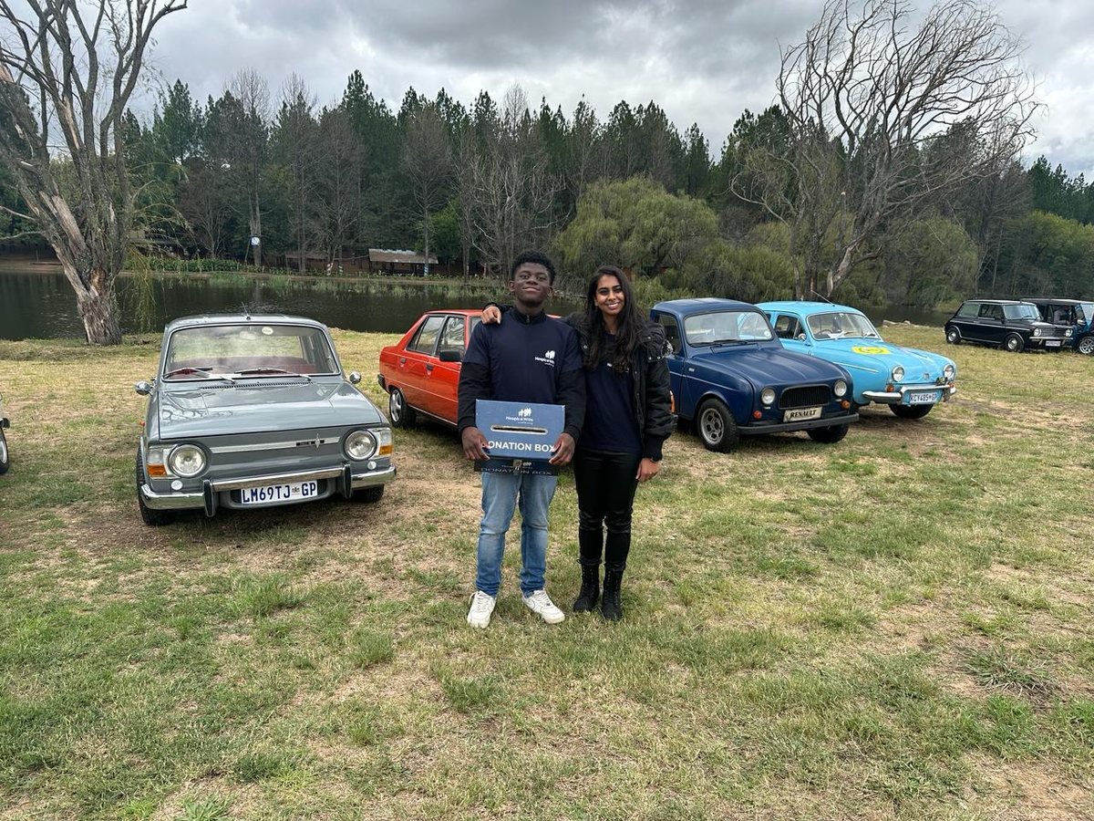 Two HospiceWits volunteers holding a donation box at a classic car show, with vintage cars and a lake behind them